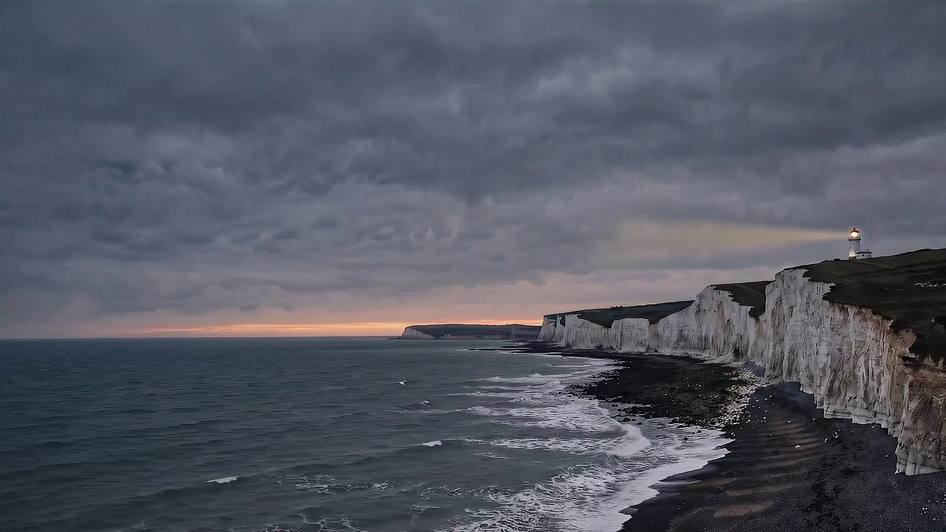 Waves breaking below the Seven Sisters at dusk, seen from Seaford Head, with Belle Tout lighthouse lit on the far clifftop